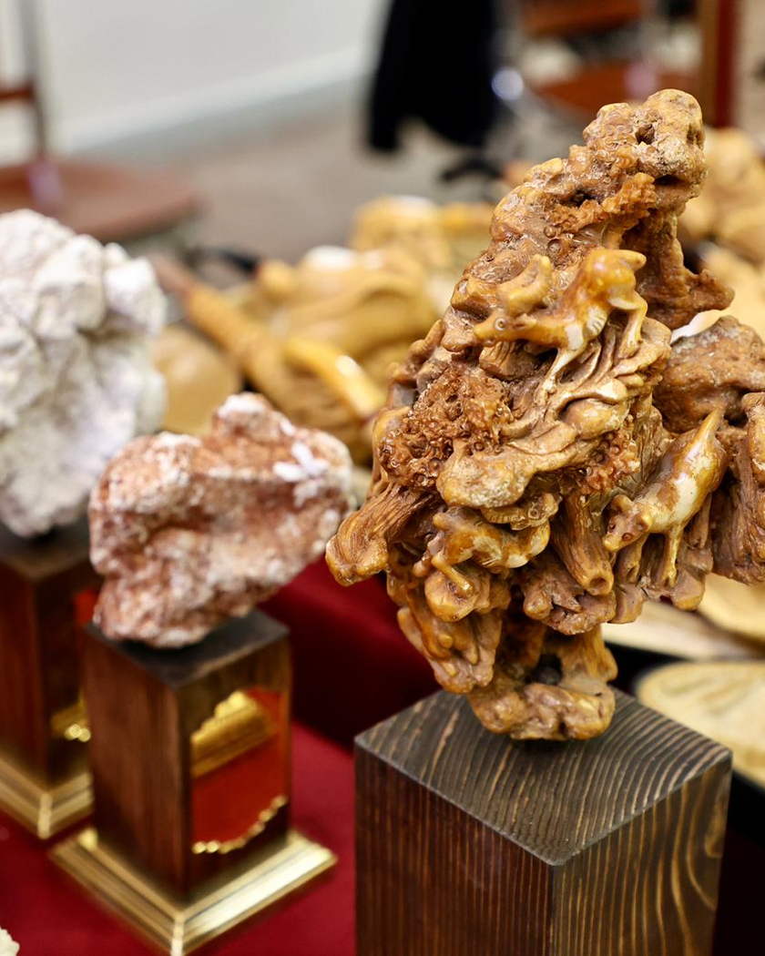 Kırmızı kumaş örtülü bir masada, ahşap kaideler üzerinde doğal lületaşı yumruları ve bal renginde oyulmuş eserler.