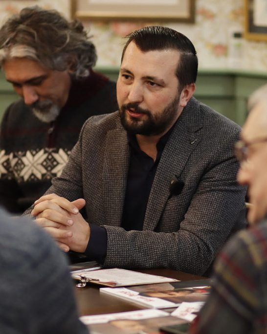 Emre Mangaltepe, ahşap bir masanın başında elleri kenetli biçimde öne eğilmiş, karşısındaki kişilerle konuşuyor; masanın üzerinde basılı yayınlar duruyor.