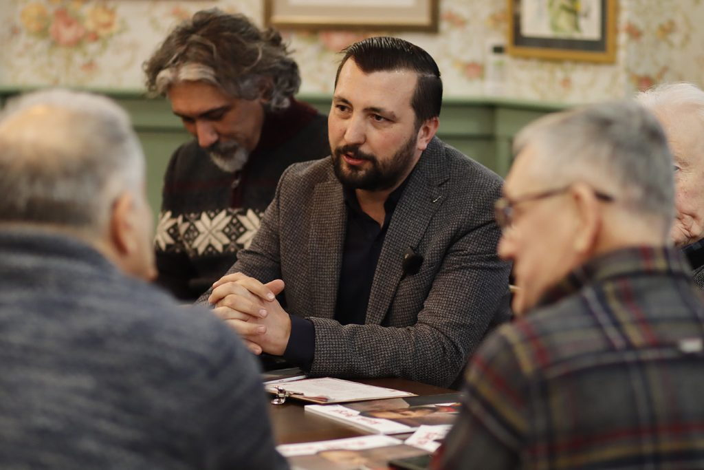 Emre Mangaltepe, ahşap bir masanın başında elleri kenetli biçimde öne eğilmiş, karşısındaki kişilerle konuşuyor; masanın üzerinde basılı yayınlar duruyor.