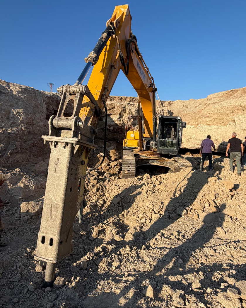 Hidrolik kırıcısı yere dayalı ekskavatör ve arkada ocak basamağında duran iki kişi.