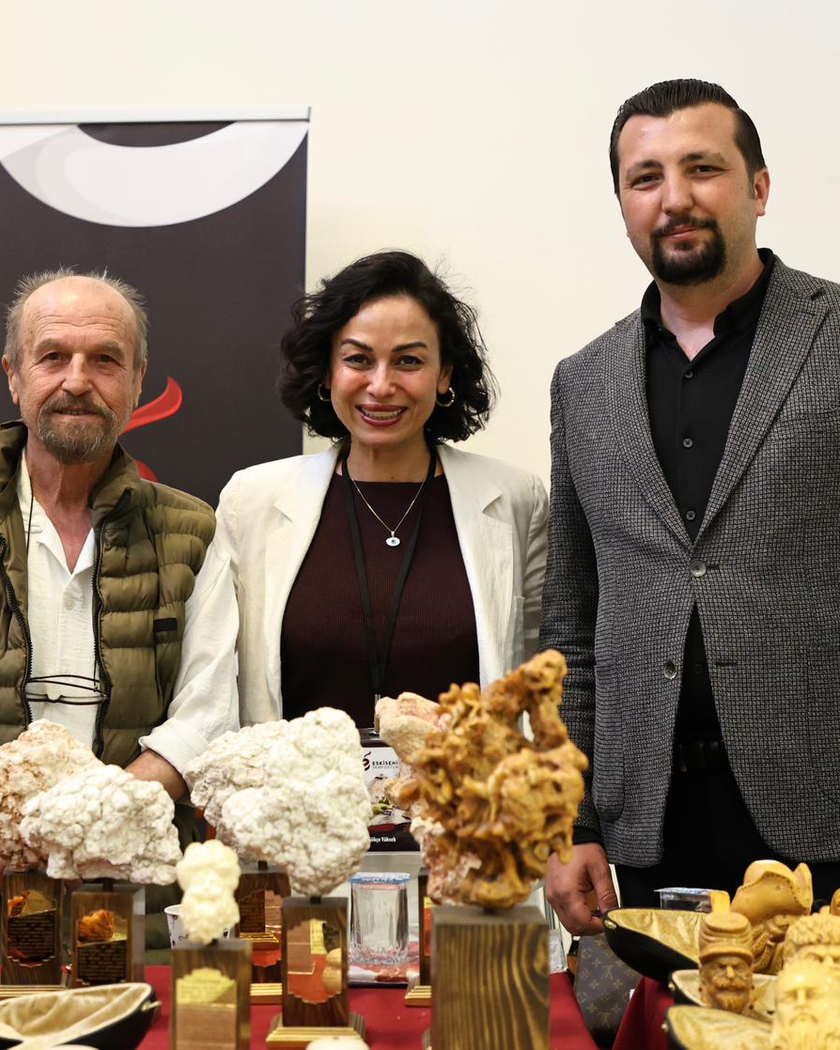 Doğal lületaşı yumruları ve oyma eserlerle dolu masanın arkasında yan yana duran üç kişi; arkada konferans salonu yönlendirme tabelası.