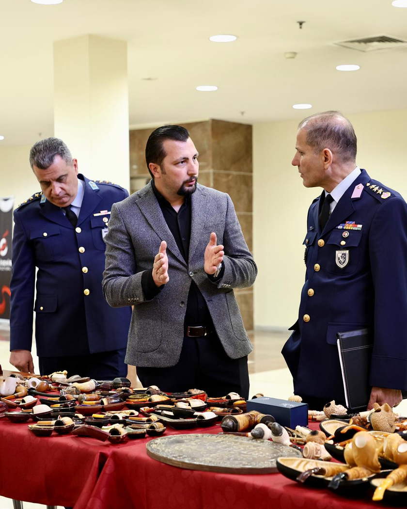 Kırmızı örtülü sergi masasının başında ayakta duran ve konuşan üç kişi.