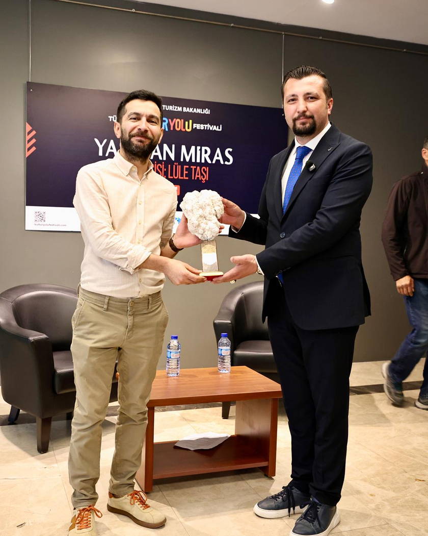 Festival afişinin önünde, birlikte bir doğal lületaşı yumrusu tutan iki kişi; afişte “Yaşayan Miras” ve “Lüle Taşı” ifadeleri okunuyor.