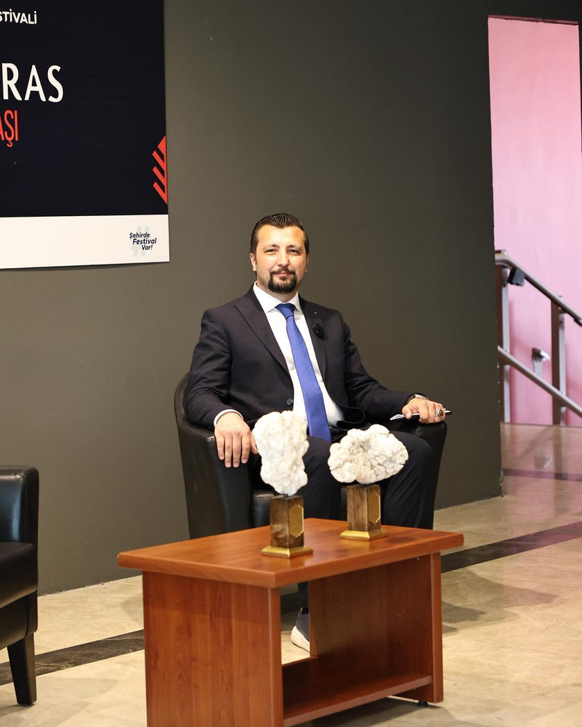 Üzerinde festival afişi bulunan duvarın önünde tek koltukta oturan bir kişi; önündeki sehpada kaideli iki doğal lületaşı örneği.