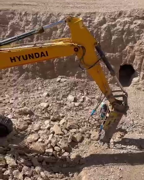 Açık ocak basamağında hidrolik kırıcı takılı bir ekskavatörün çalışması ve ocak alanının panoramik görüntüsü.